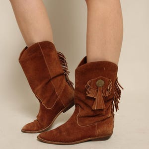 May include: A pair of mid-calf brown suede boots featuring a pointed toe, a flat sole, and decorative fringe detailing along the outer side. Each boot is accented with a circular metal concho ornament with hanging fringe tassels on the side.