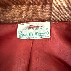 以下が含まれることがあります： ヴィンテージコートの内側のクローズアップ。赤い裏地と「Sears Jr. Bazaar」のラベル、「ドライクリーニング」の指示が表示されています。コートの外側は茶色と赤のチェック柄です。