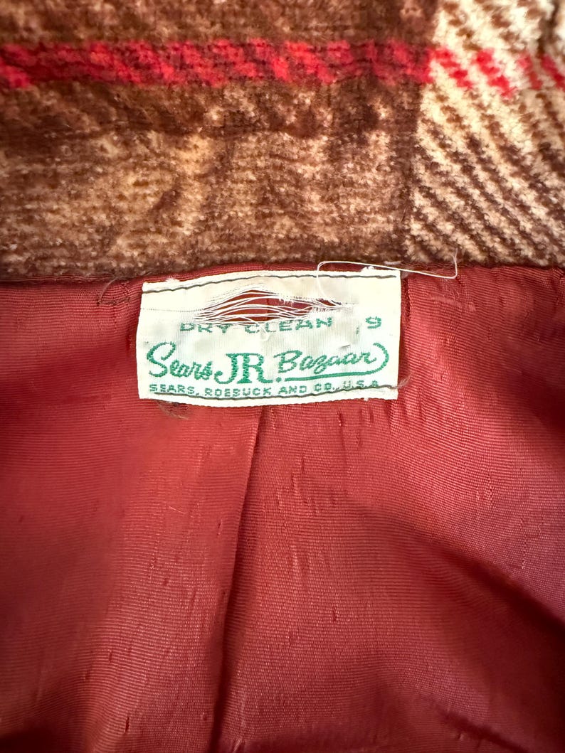 以下が含まれることがあります： ヴィンテージコートの内側のクローズアップ。赤い裏地と「Sears Jr. Bazaar」のラベル、「ドライクリーニング」の指示が表示されています。コートの外側は茶色と赤のチェック柄です。