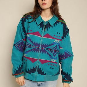 May include: A teal zip-up jacket featuring a bold geometric pattern in shades of dark blue, magenta, and grey. The jacket has a classic collar, long sleeves with ribbed cuffs, and a matching ribbed hem, constructed from a thick, textured fabric.