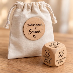 Könnte beinhalten: Ein beigefarbener Stoffbeutel mit Kordelzug und einem runden Holzanhänger mit der Aufschrift Gefühlszeit mit Emma und einem Herzsymbol. Daneben liegt ein Holzwürfel, der mit deutschem Text und Symbolen wie Sonne und Gesichtern graviert ist.