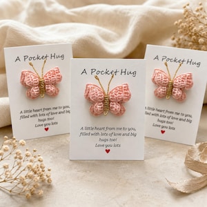 Könnte beinhalten: Drei weiße rechteckige Karten mit jeweils einem gehäkelten rosa Schmetterling mit goldenen Drahtantennen. Der Text auf jeder Karte lautet A Pocket Hug, A little heart from me to you, filled with lots of love and big hugs too! Love you lots, mit einem roten Herz.