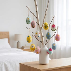 May include: A birch branch with hanging felt Easter egg ornaments. The eggs are in various pastel colors, decorated with embroidered flowers and dots. The branch is in a white vase on a wooden table.