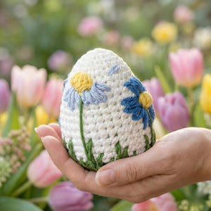 K&ouml;nnte beinhalten: Ein wei&szlig;es geh&auml;keltes Ei, verziert mit blauen und gelben G&auml;nsebl&uuml;mchen und gr&uuml;nen Stielen. Das Ei wird in einer Hand gehalten, mit einem verschwommenen Hintergrund aus bunten Blumen.