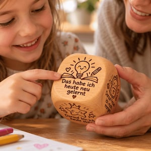 Könnte beinhalten: Ein heller Holzwürfel mit lasergravierten Illustrationen und deutschem Text. Die Vorderseite zeigt ein offenes Buch, eine Glühbirne und einen Stift mit dem Text 'Das habe ich heute neu gelernt'. Weitere Seiten zeigen einen Rucksack und einen Stern.