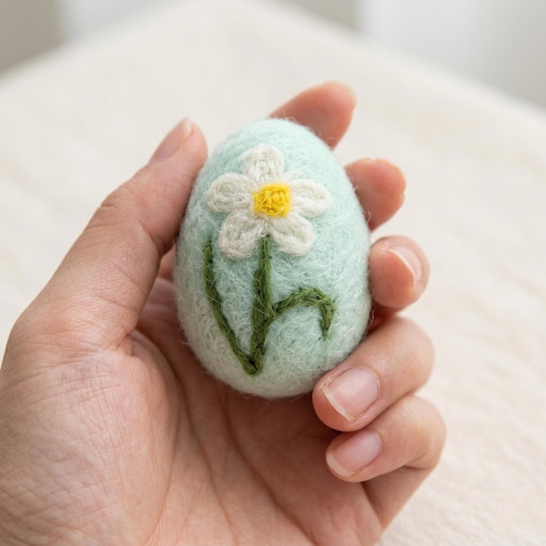 May include: A light blue felt Easter egg with a white and yellow flower design. The egg is hand-stitched with green embroidery for the stem and leaves. The egg is held in a hand.