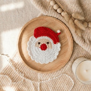 Könnte beinhalten: Ein runder, gehäkelter Untersetzer in Form eines Weihnachtsmann-Gesichts mit roter Mütze und weißem Bommel, beigem Gesicht, leuchtend roter Nase, zwei kleinen schwarzen Augen und einem strukturierten weißen Bart auf einem runden Holzteller.