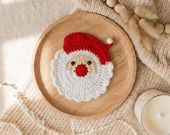 May include: A round, crocheted Santa Claus face coaster featuring a red hat with a white pom-pom, a beige face, a bright red nose, two small black eyes, and a textured white beard. The coaster is centered on a smooth, circular wooden plate.
