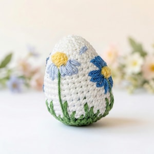 K&ouml;nnte beinhalten: Ein wei&szlig;es, geh&auml;keltes, eif&ouml;rmiges Ornament mit gestickten blauen und gelben G&auml;nsebl&uuml;mchen und gr&uuml;nem Gras am Boden. Die handgefertigte Dekoration eignet sich f&uuml;r Ostern oder Fr&uuml;hlingsdekoration.