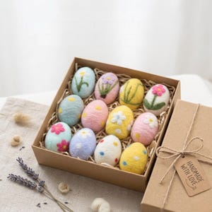 Puede incluir: Una docena de huevos de Pascua de fieltro de colores con diseños florales en una caja de cartón marrón. Los huevos son de tonos pastel. Una etiqueta en la caja dice "HANDMADE WITH LOVE".