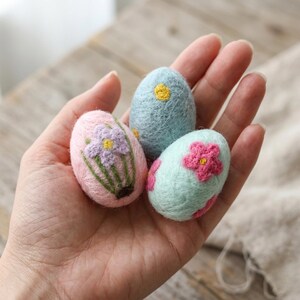May include: Three colorful, embroidered Easter eggs. One is pink with a purple flower, another is light blue with a yellow flower, and the third is mint green with pink flowers. The eggs are held in a hand.