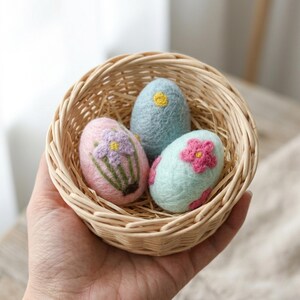 May include: A hand holds a small woven basket filled with three decorative Easter eggs. The eggs are pastel pink, blue, and mint green, each adorned with embroidered floral designs in shades of pink, purple, and yellow. The basket is filled with straw.