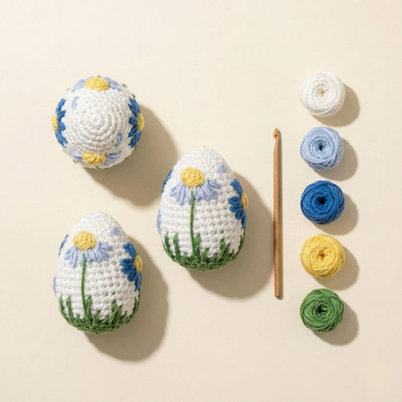 K&ouml;nnte beinhalten: Wei&szlig;e geh&auml;kelte Ostereier mit blauen und gelben Blumendetails und gr&uuml;nen Grasbasen. Ein H&auml;kelhaken aus Holz und mehrere Kn&auml;uel Garn in Wei&szlig;, Blau, Gelb und Gr&uuml;n sind ebenfalls vorhanden. Die Artikel sind auf einer cremefarbenen Oberfl&auml;che angeordnet.