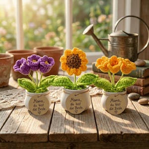 Puede incluir: Tres arreglos florales de ganchillo en macetas blancas con el texto "Mama, du bist die Beste." Cada maceta tiene una flor diferente: morada, amarilla y naranja. Las flores están sobre una superficie de madera.
