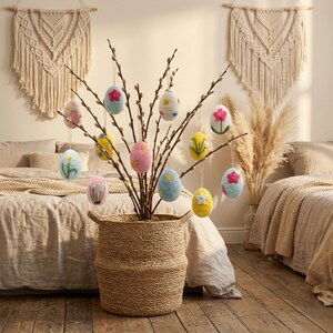 May include: A woven basket holds branches adorned with colorful, felt Easter egg ornaments. The eggs are decorated with floral designs in shades of pink, blue, yellow, and white. The basket sits on a wooden floor, with macrame wall hangings in the background.