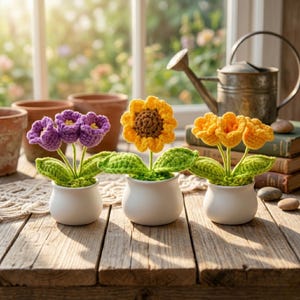 Può includere: Tre composizioni floreali all'uncinetto in vasi bianchi su un tavolo di legno. I fiori sono viola, gialli e arancioni, con foglie verdi. Un annaffiatoio e dei libri sono sullo sfondo, vicino a una finestra.