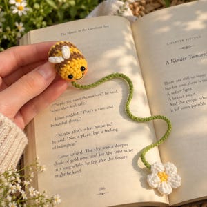 Può includere: Un segnalibro all'uncinetto con una piccola ape rotonda a strisce gialle e marroni con ali bianche, attaccata a un lungo e sottile stelo di filo verde che termina con un fiore bianco dal centro giallo, appoggiato su una pagina di libro.