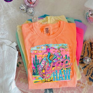 Könnte beinhalten: Ein Stapel bunter T-Shirts, mit einem orangefarbenen Shirt oben, das eine Grafik eines Rodeo-Reiters und das Wort "HAW" zeigt. Die Shirts sind in Orange-, Gelb-, Grün-, Rosa- und Lilatönen gehalten. Ein weißer Cowboy-Stiefel ist links zu sehen.