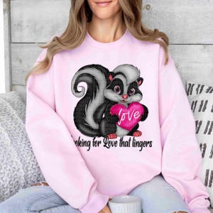 May include: Pink sweatshirt with a graphic of a cartoon skunk holding a pink heart with the word "love" on it. The text "Looking for love that lingers" is printed below the skunk.