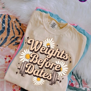 May include: A stack of three t-shirts in pastel colors: light yellow, pink, and blue. The top shirt is light yellow with the text "Weights Before Dates" in a retro font, decorated with daisies and a barbell graphic.