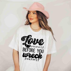 May include: White t-shirt with black text that reads "Love yourself before you wreck yourself". The text is in a retro font style. A woman is wearing the shirt and a pink cowboy hat.