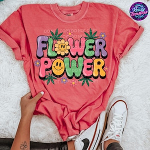 May include: A coral-colored t-shirt with the words "FLOWER POWER" in a colorful, retro font. The design includes a smiling flower, flowers, and cannabis leaves. The shirt is paired with white sneakers.