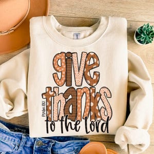 May include: A cream-colored sweatshirt with the words "give thanks to the Lord" in brown and orange letters with a polka dot pattern. The text "Psalm 100:4" is printed in small letters below the main text.
