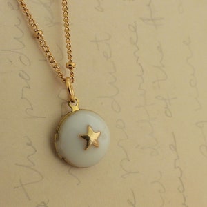 May include: A gold chain necklace with a small round locket pendant. The locket is white with a gold star in the center.