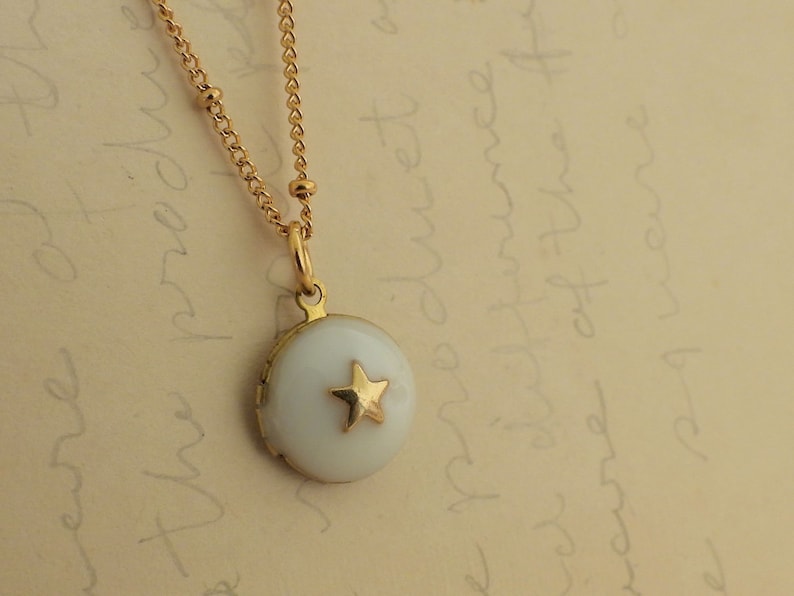 May include: A gold chain necklace with a small round locket pendant. The locket is white with a gold star in the center.