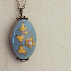 May include: A blue oval locket necklace with a delicate floral design in yellow and white. The locket features a small pearl in the center of the flower.