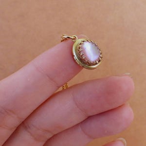 Könnte beinhalten: Eine goldene Halskette mit einem Anhänger in Form eines Medaillons, der mit einem rosa schimmernden Stein in einem goldenen Filigranrahmen besetzt ist.