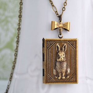 May include: A gold locket necklace featuring a small bow charm and a detailed image of a rabbit inside. The locket is attached to a gold chain.