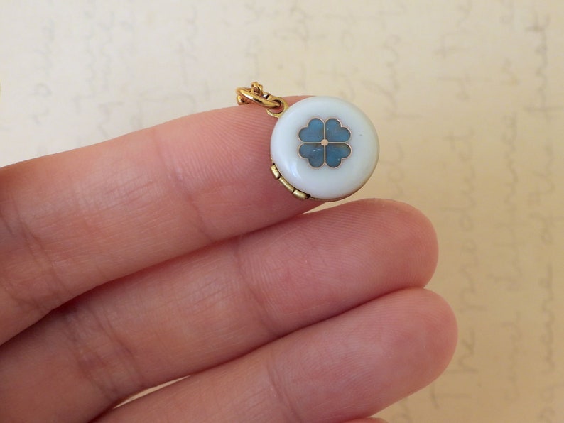 May include: A gold locket pendant with a white enamel round charm featuring a blue four-leaf clover design.