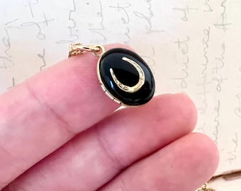 Op de afbeelding: Een goudkleurige ketting met een ovale, zwarte hanger met een gouden hoefijzerontwerp. De hanger wordt door een hand vastgehouden, waarbij de details van de sieraden tegen een zachte achtergrond worden getoond.