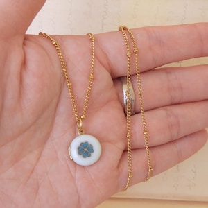 May include: A gold chain necklace with a small round locket pendant. The locket is white with a blue four-leaf clover design.