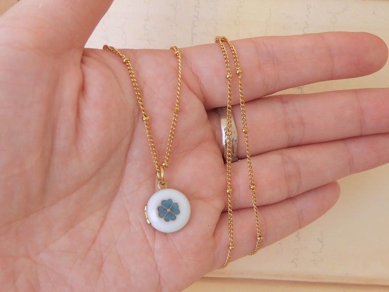 May include: A gold chain necklace with a small round locket pendant. The locket is white with a blue four-leaf clover design.