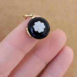 May include: A gold locket necklace with a black enamel round pendant featuring a white flower with a gold center.