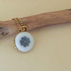 May include: A gold chain necklace with a white locket pendant featuring a blue four-leaf clover design.