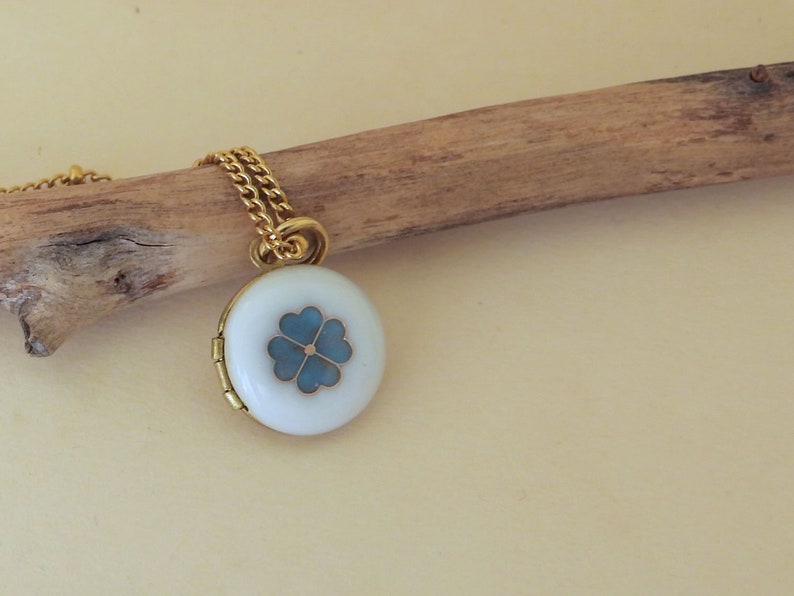 May include: A gold chain necklace with a white locket pendant featuring a blue four-leaf clover design.