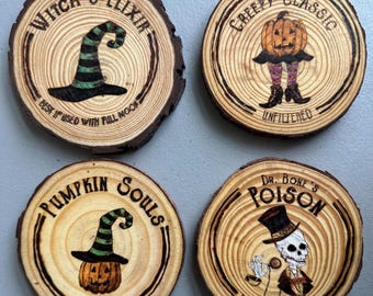 Op de afbeelding: Vier houten onderzetters met Halloween-ontwerpen. Eén onderzetter leest "Witch's Elixir" met een heksenhoed. Een andere zegt "Children's Creepy Classic" met een pompoen. De derde zegt "Pumpkin Souls" met een pompoen en heksenhoed. De laatste zegt "Dr. Bone's Poison".