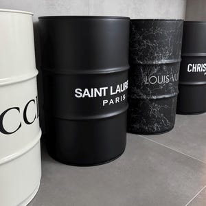 Può includere: Una fila di quattro fusti metallici cilindrici. Il primo è bianco con testo nero GUCCI. Il secondo è nero opaco con testo bianco SAINT LAURENT PARIS. Il terzo è nero con un motivo a marmo bianco e testo LOUIS VUITTON. Il quarto è nero con testo bianco CHRISTIAN DIOR PARIS.