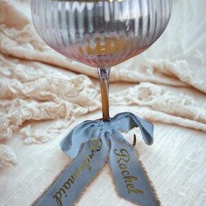 May include: A champagne glass with a gold rim and a light blue ribbon tied around the stem. The ribbon has the words "Bridesmaid" and "Rachel" printed in gold lettering. The glass has vertical ridges.