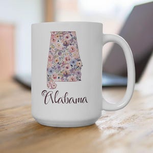May include: White ceramic mug featuring the state of Alabama filled with a floral pattern in shades of pink, purple, and blue. The word "Alabama" is written in cursive below the state outline.