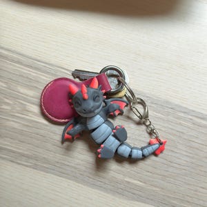 May include: A gray and red dragon keychain with a silver keyring and a burgundy leather tag. The dragon has red horns, wings, and accents. The keychain is lying on a light wood surface.