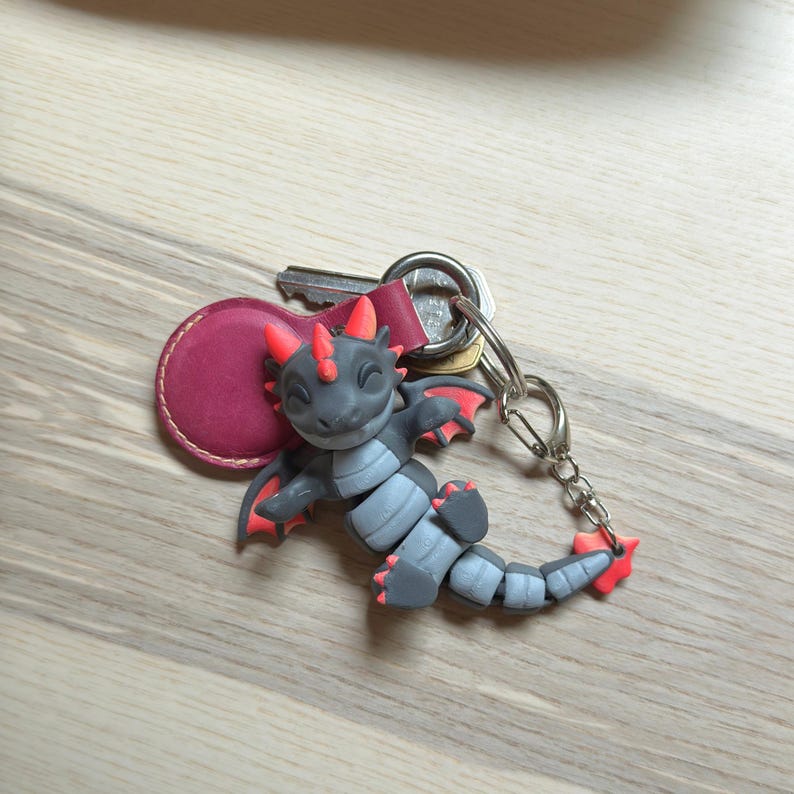 May include: A gray and red dragon keychain with a silver keyring and a burgundy leather tag. The dragon has red horns, wings, and accents. The keychain is lying on a light wood surface.
