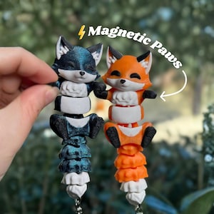 May include: Two fox-shaped magnetic toys. One is blue and black, the other is orange and white. The toys have articulated bodies and are connected to a keychain. The text "Magnetic Paws" is in the image.