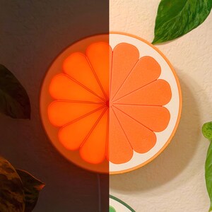 Orange slice wall lamp shown lit and unlit in one frame — glowing warm tangerine on one half, true matte orange on the other. Dimensional retro fruit wall art and a cute night light for a dopamine decor room or reading nook.