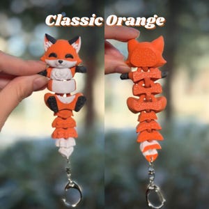 Classic Orange 3D printed flexi fox keychain shown front and back. Front view shows the smiling face, articulated body segments, and white belly. Back view reveals the full segmented spine running down to a striped ringed tail with silver lobster clasp. Bold orange with dark grey-black accents. Classic orange fox keychain 3D printed fidget articulated bag charm backpack charm fox lover gift