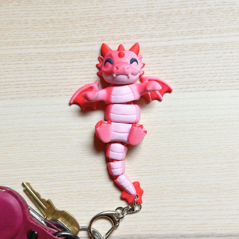 May include: A pink and red dragon keychain with a silver clasp. The dragon has a smiling face, small wings, and a segmented body. The keychain is attached to a pink leather key holder with keys.