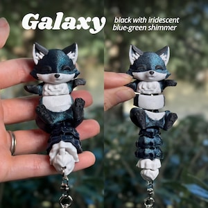 Galaxy colorway 3D printed flexi fox keychain from two angles. Deep black base with a mesmerizing iridescent blue-green shimmer that shifts and glows at every angle. Fully segmented articulated body with white facial markings and white belly. Galaxy fox keychain dark 3D printed fidget bag charm blue shimmer black fox articulated backpack charm fidget toy fox lover gift cute animal keychain